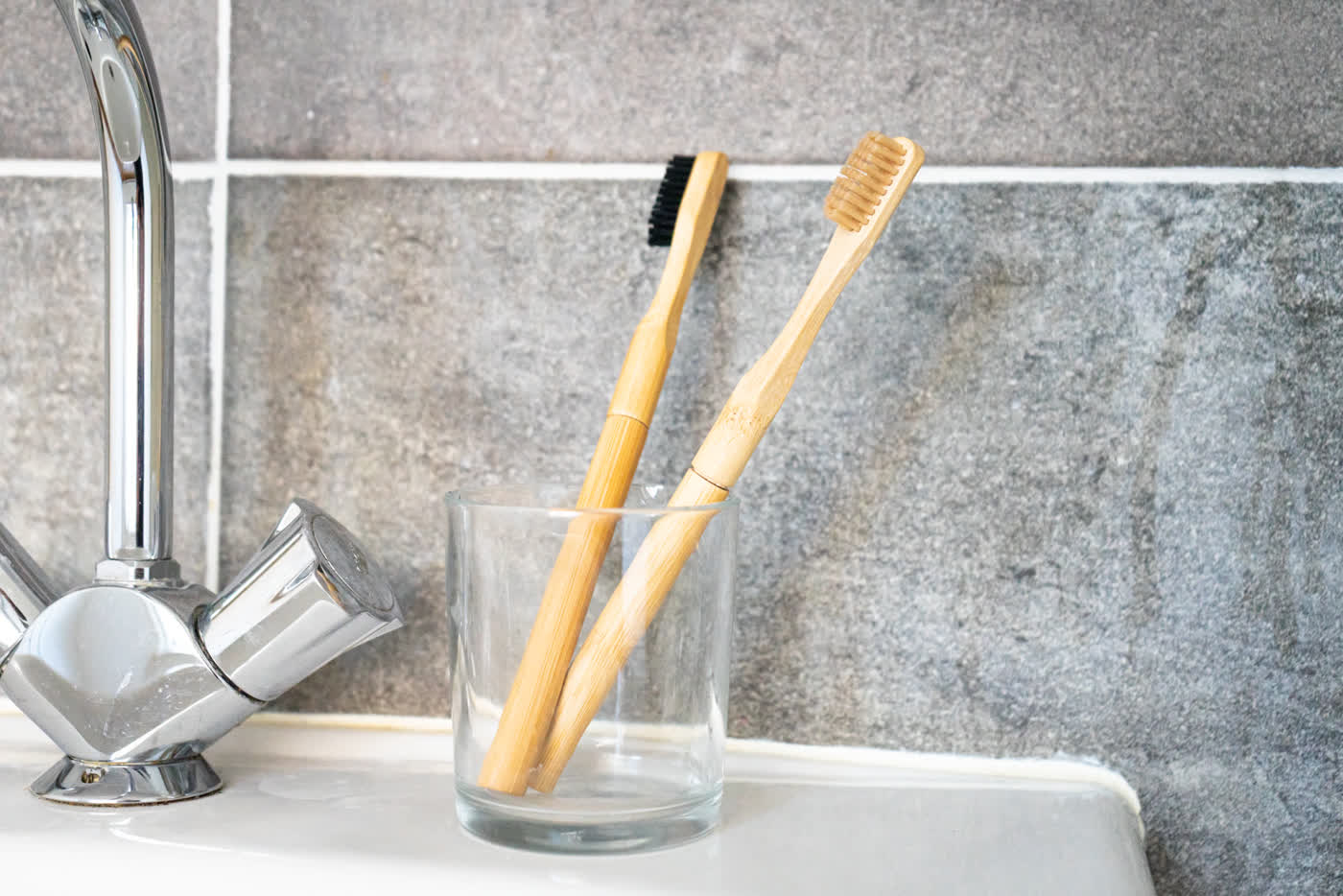 Two bamboo toothbrushes in a glass beside a chrome tap on a tiled wall